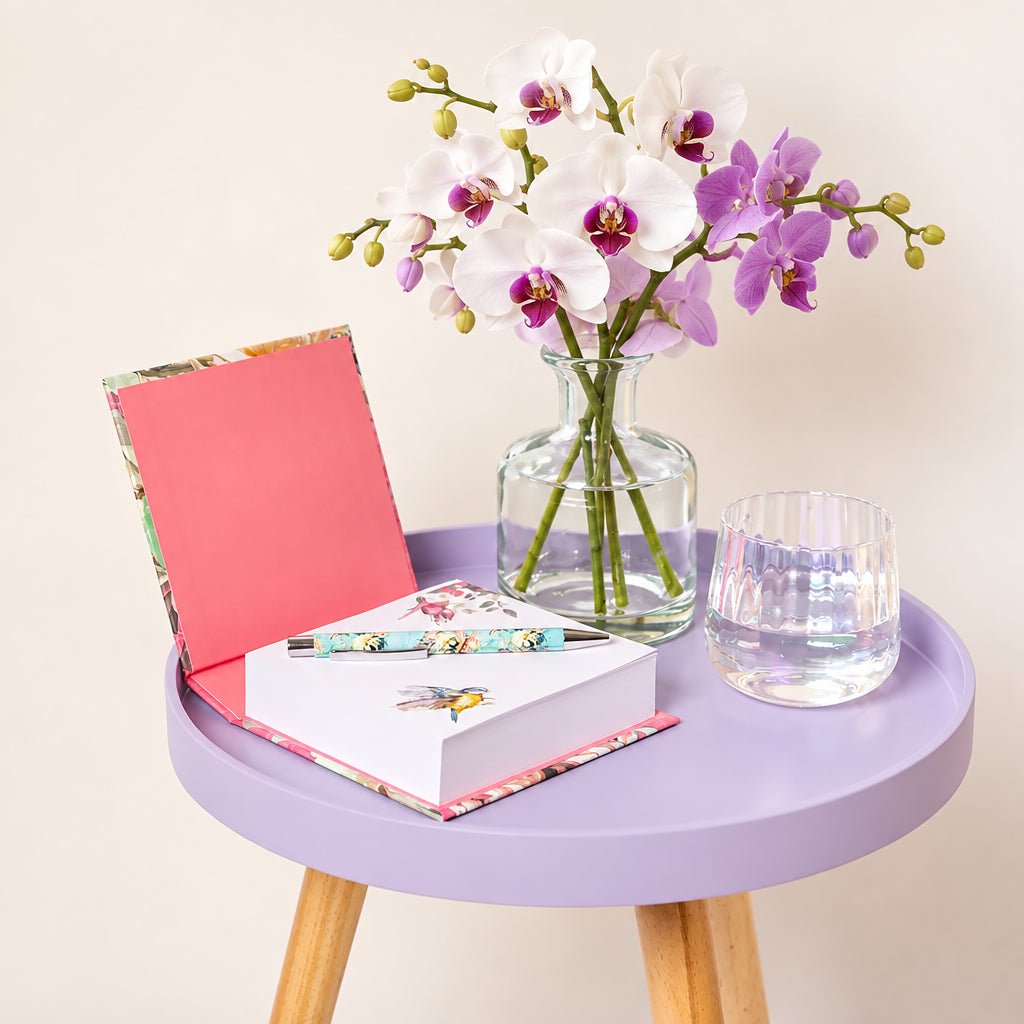 A styled tabletop scene with a round pale blue side table holding a pink notebook, a white notepad with a floral pen, a clear glass vase with eucalyptus stems, and a clear drinking glass against a plain light beige wall.