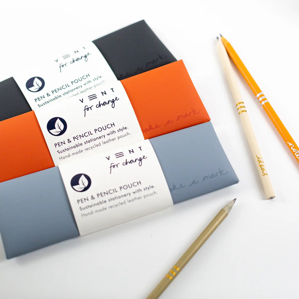 Three rectangular pen and pencil pouches wrapped in white paper bands are arranged on a white surface, alongside two pencils. The pouches are dark gray, bright orange, and light blue-gray.