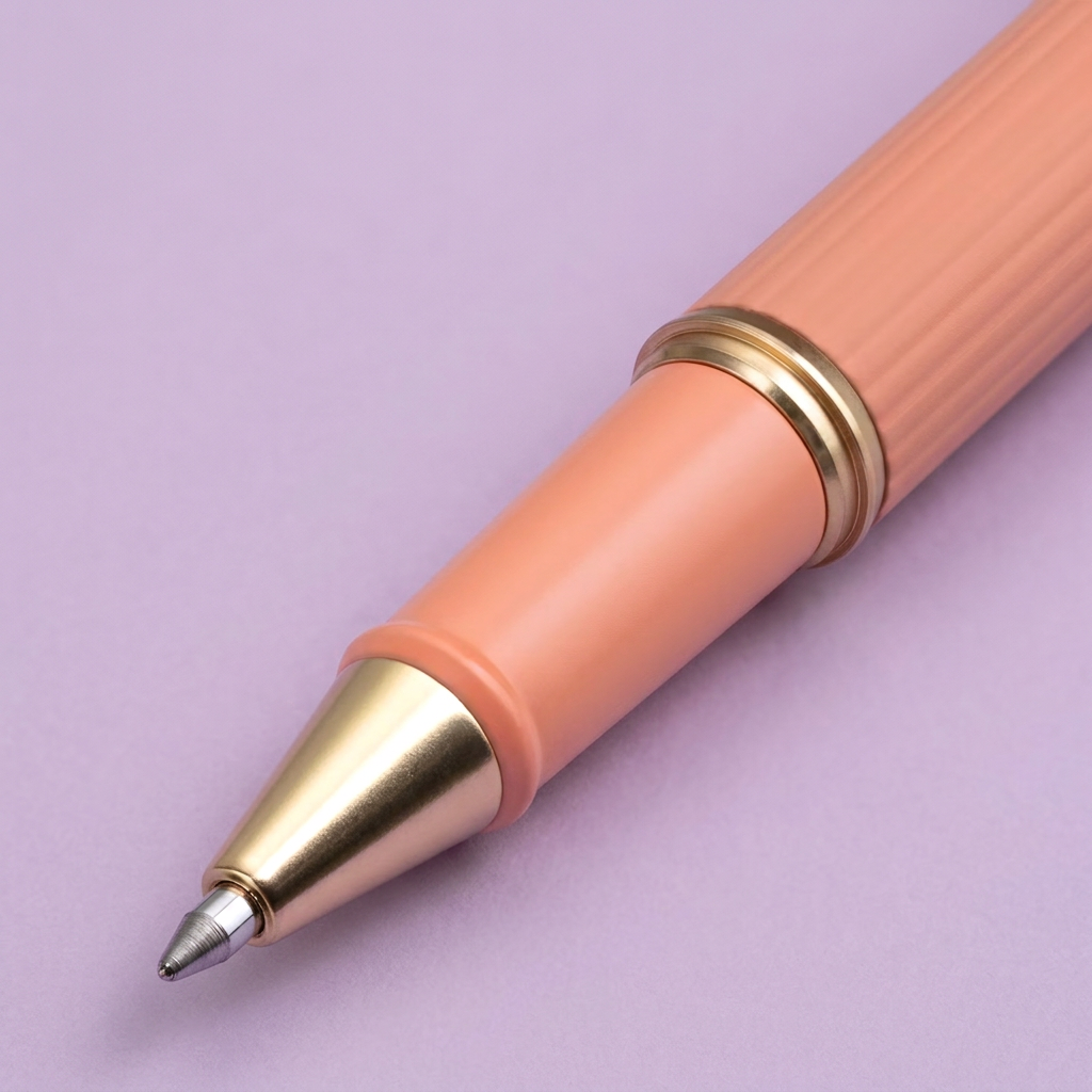 A close-up of a peach pink pen with gold metal accents, including a gold band and a gold tip section, on a clean lilac background.