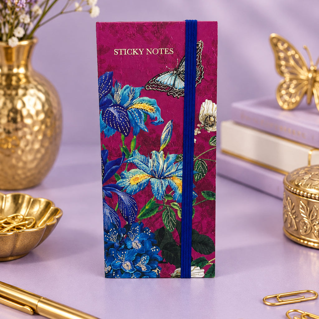 A vertical notebook of sticky notes with a deep magenta floral cover, featuring blue and yellow flowers, a butterfly, and a blue elastic closure band, set against a neutral gray background.