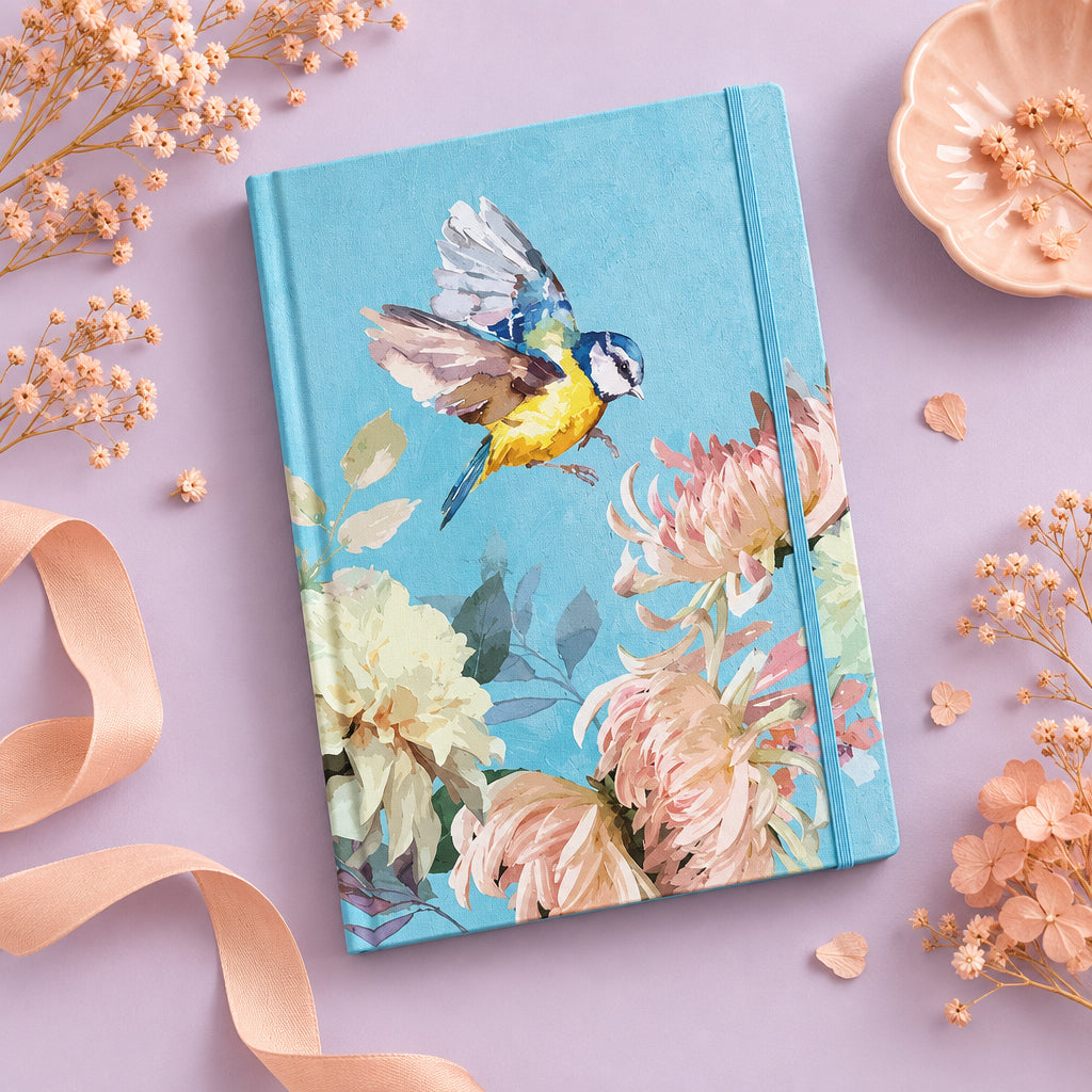 A hardcover notebook with a vibrant blue cover featuring a painted bluebird in flight and pink and cream flowers, shown at an angle on a light gray background.