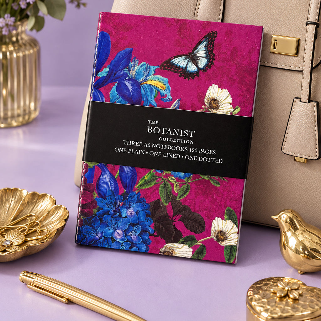 Botanist Collection A6 notebook set in its black packaging band, showing a magenta floral and butterfly cover, leaning against a cream handbag on a lilac surface