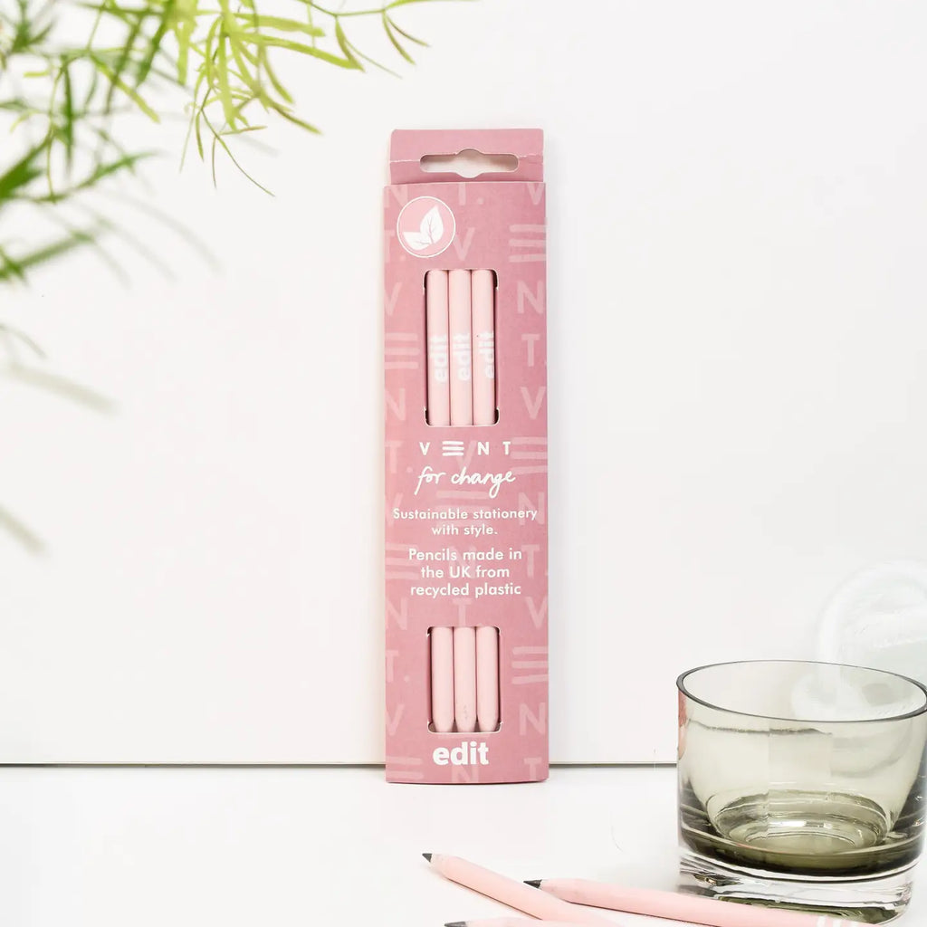 Three powder pink recycled pencils in a matching pink presentation sleeve, beside a plant and a glass.