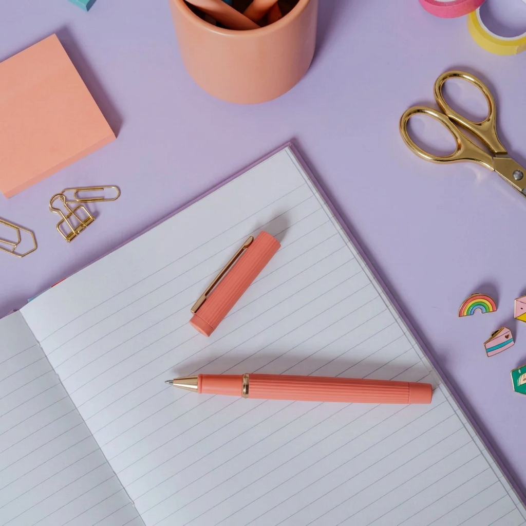 A peach ribbed fountain pen with gold trim lies on an open lined notebook on a light wood desk, with the pen cap placed nearby. Gold scissors and clips and peach pen holder and notepad  next to it