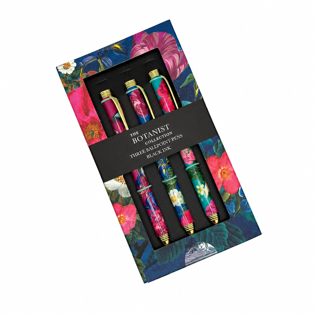 The Botanist Collection three ballpoint pens in a floral-patterned gift box with a black paper band. The pens feature vibrant pink, green, and blue botanical designs with gold accents.