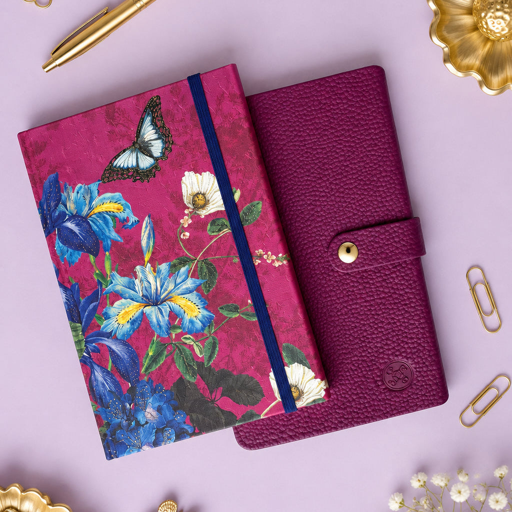 Two notebooks on a light-colored tabletop. One notebook has a textured magenta cover with a silver snap button, and the other has a vibrant floral and butterfly pattern on a magenta background with a blue elastic closure.