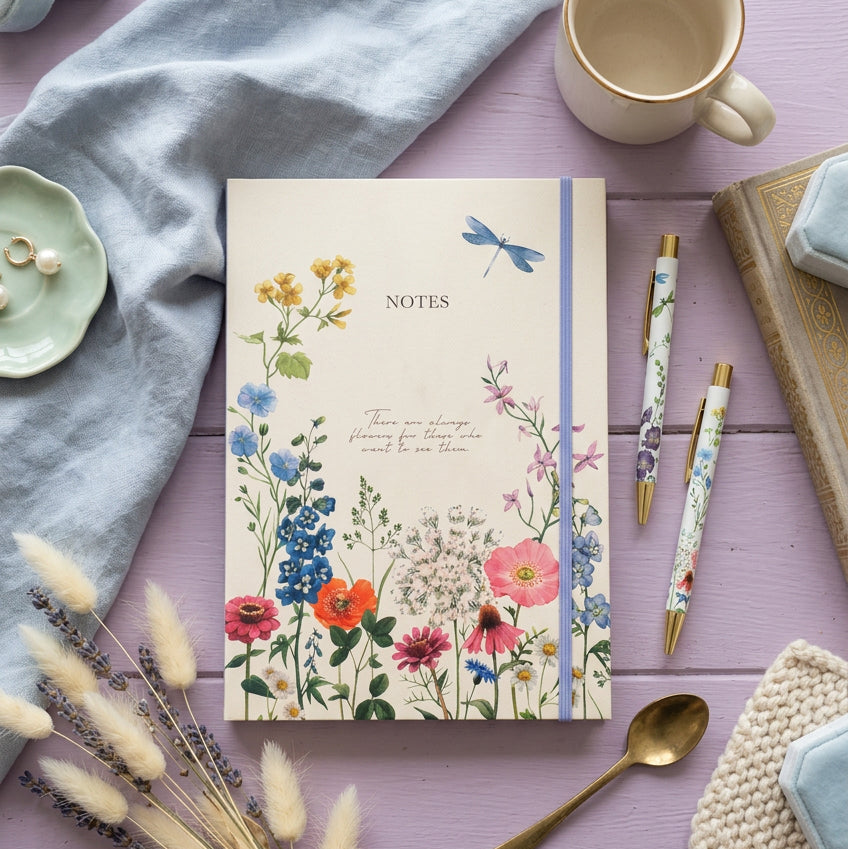 A cream-colored notebook with a blue elastic band and floral illustrations, shown on a lilac surface next to two gold pen-style pencils.