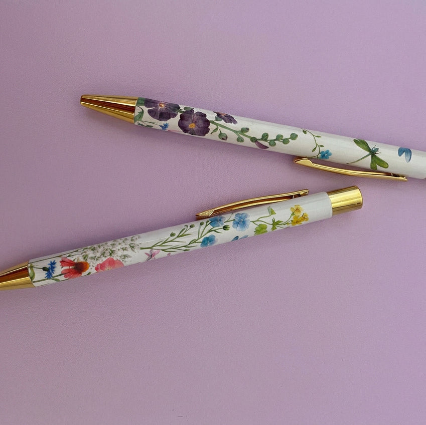 Two ballpoint pens with gold metal tips and clips, featuring a white barrel decorated with delicate multicolor floral illustrations, displayed on a lilac background.