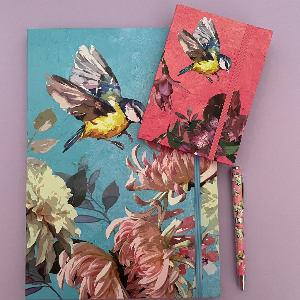 Two floral and bird illustrated notebooks or journals are arranged on a lilac background, with one larger blue cover and one smaller pink cover, plus a matching patterned pen.