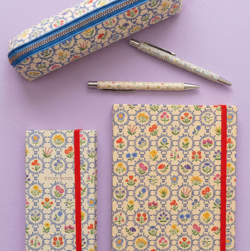 A coordinated stationery set laid out on a light gray surface, including a zippered pencil case, two pens, and two notebooks with a repeating floral pattern. The set features a lilac background with multicolored floral motifs and red elastic closure bands.