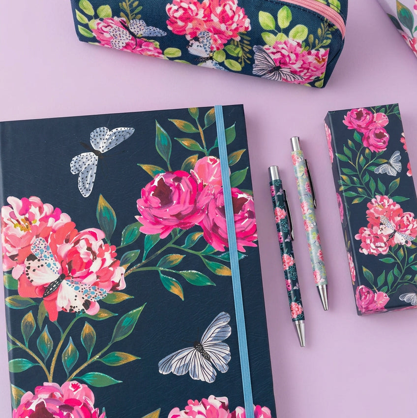 A floral stationery set on a lilac desk, featuring a notebook with a dark navy background, pink roses, green leaves, and white butterflies, alongside a matching pen, pencil, and pencil case.