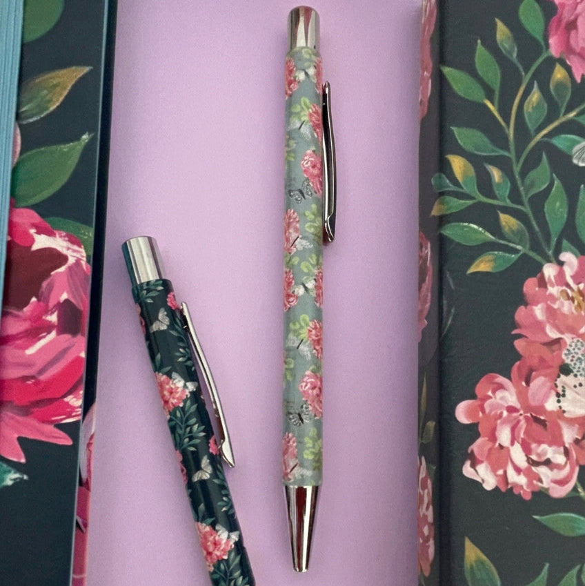Two floral metal pens with silver accents are displayed on a lilac surface next to a matching floral gift box. One pen has a pink and red floral pattern on a gray background, and the other has a pink and red floral pattern on a dark green background.