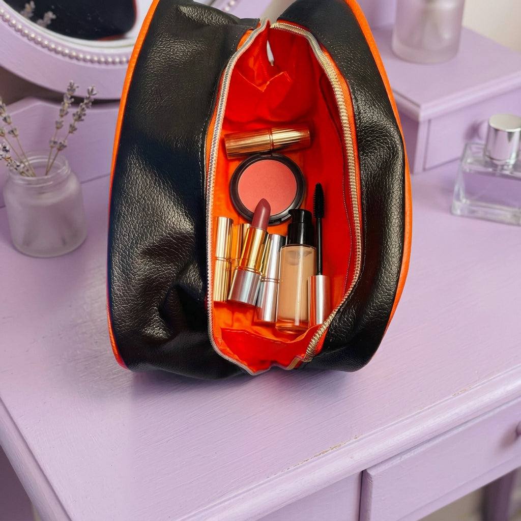 A black faux-leather zippered pouch with a bright orange interior lining, shown open with make up accessories inside against a lilac dressing table witih clear perfume bottle 