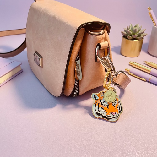 A blush pink leather shoulder bag with a gold-tone clasp, shown on a desk with a tiger-shaped enamel charm hanging from the zipper.