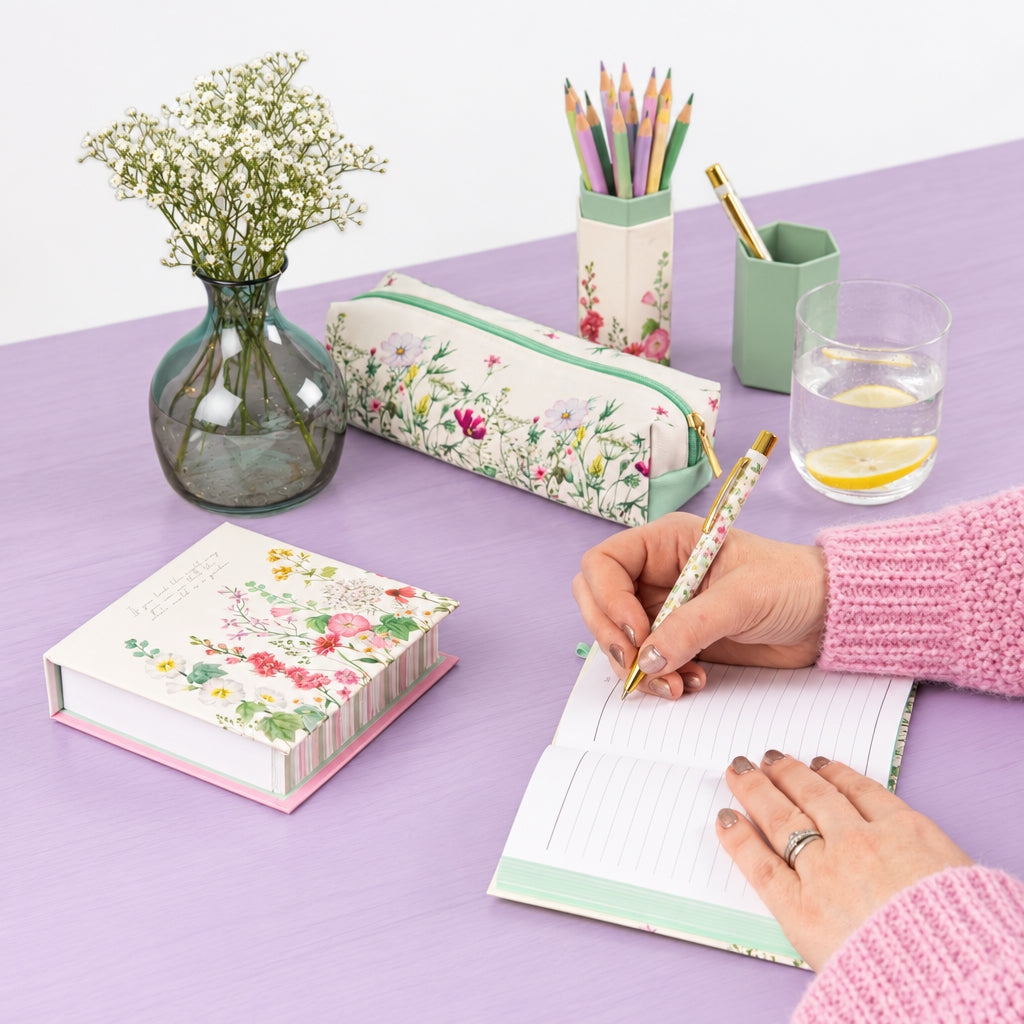 Writing in an open Wild Harmony A6 notebook with a floral pen on a lilac desk, beside a matching pencil case, floral stationery and a vase of white flowers