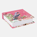 A hardcover notebook or journal with a pink cover decorated with a bluebird perched among white flowers, shown at an angle on a light gray background.
