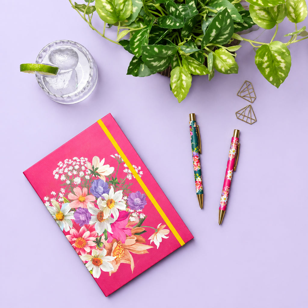 A styled flat-lay on a wooden desk featuring a bright pink floral notebook with a yellow elastic band, two matching floral pens, a small glass of water with a lime wedge, and a green plant.