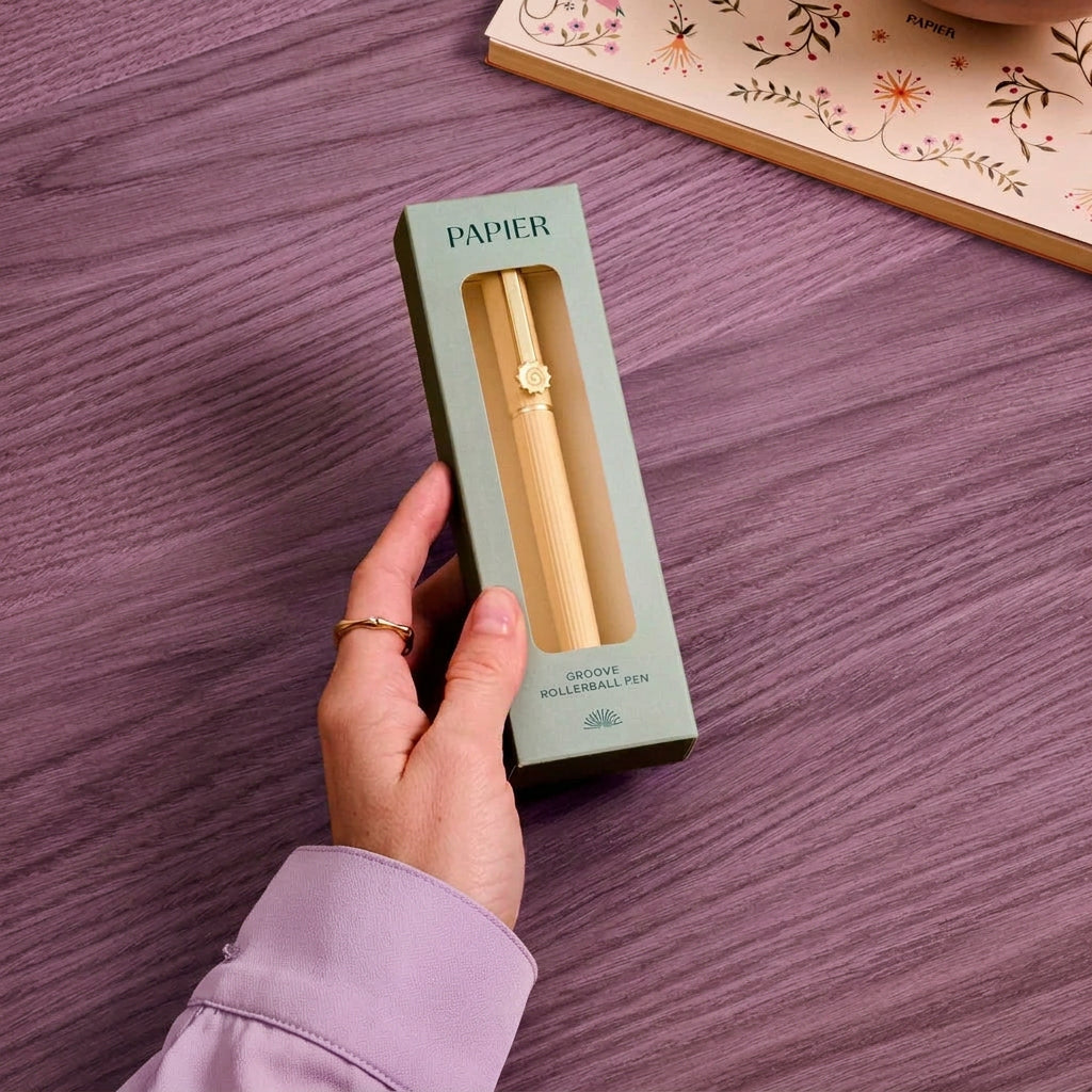 A hand holds a light blue Papiér box containing a gold rollerball pen, placed on a lilac wooden table with a floral notebook in the background.