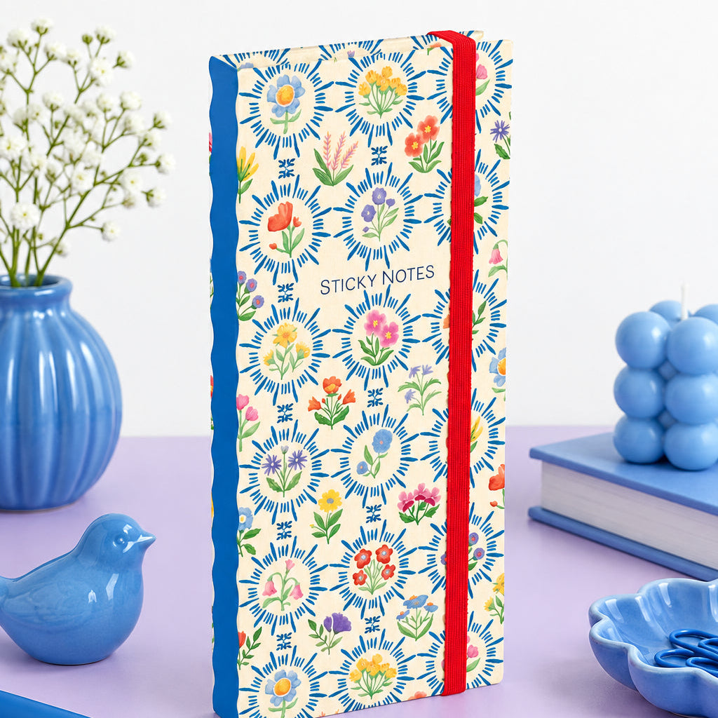 A notebook of sticky notes with a cream-colored cover decorated with a colorful floral pattern, featuring a blue spine and a red elastic closure band.