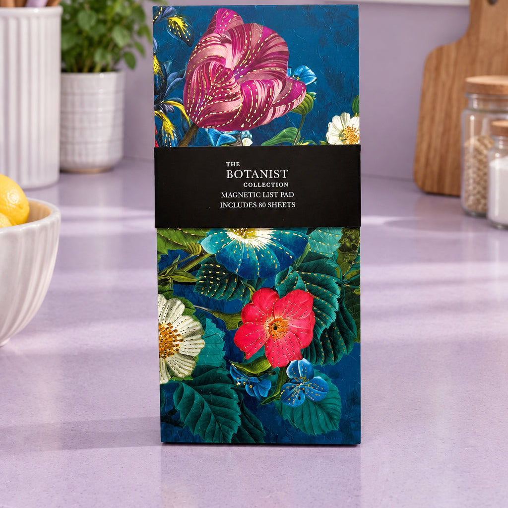 The Botanist Collection Magnetic List Pad with an illustrated botanical cover featuring pink and red flowers on a dark blue background, wrapped with a black paper band.