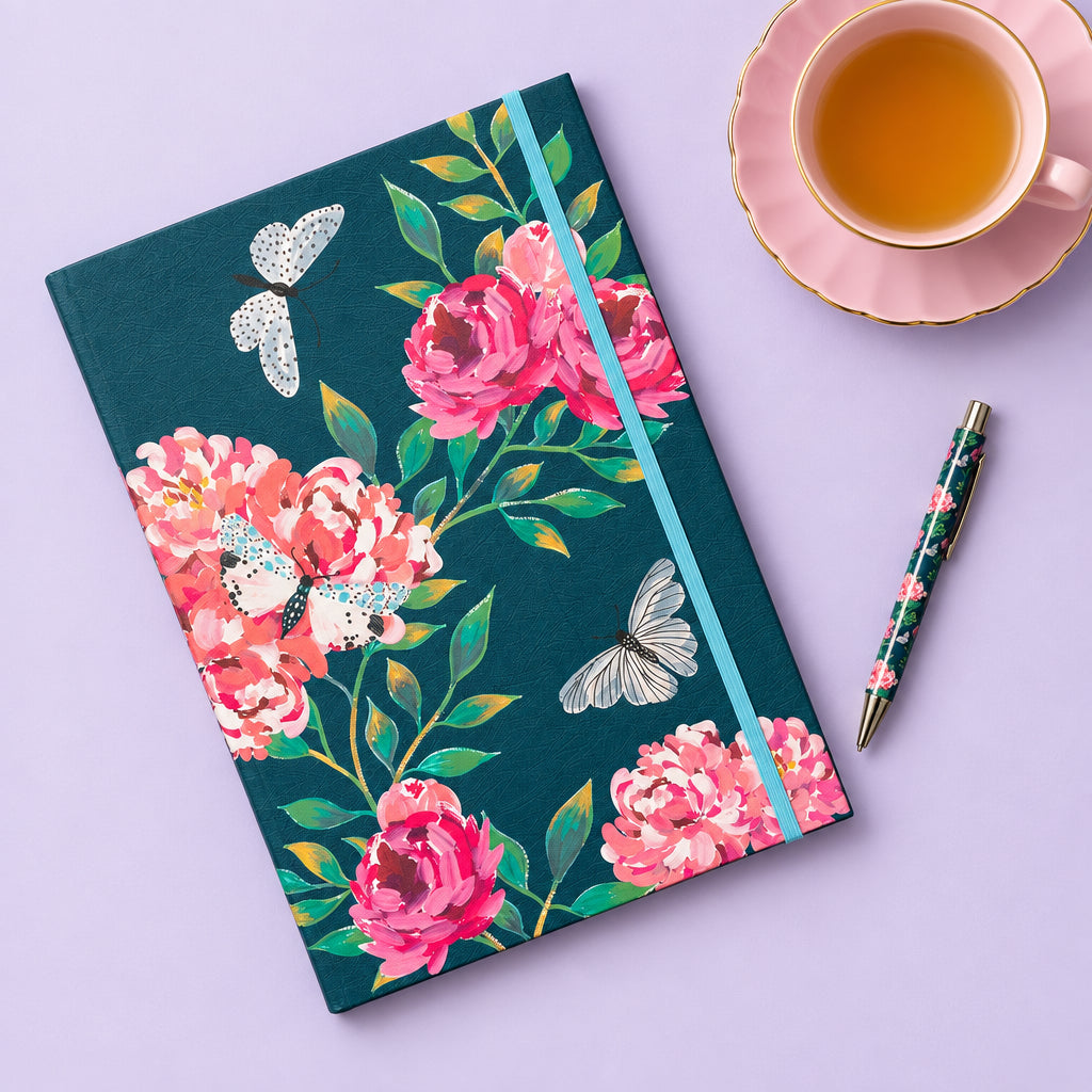 A floral notebook with a dark teal cover, pink peonies, green leaves, and white butterflies, styled on a wooden table next to a glass of lemon tea and a matching pen.