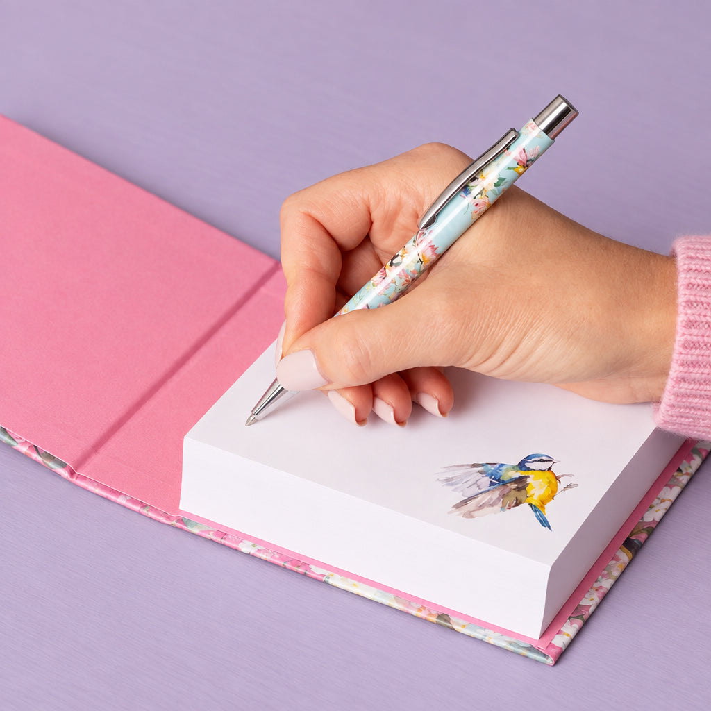 A hand holding a floral-patterned retractable pen and writing on a white notepad with a bird illustration, placed on a pink notebook over a wooden surface.
