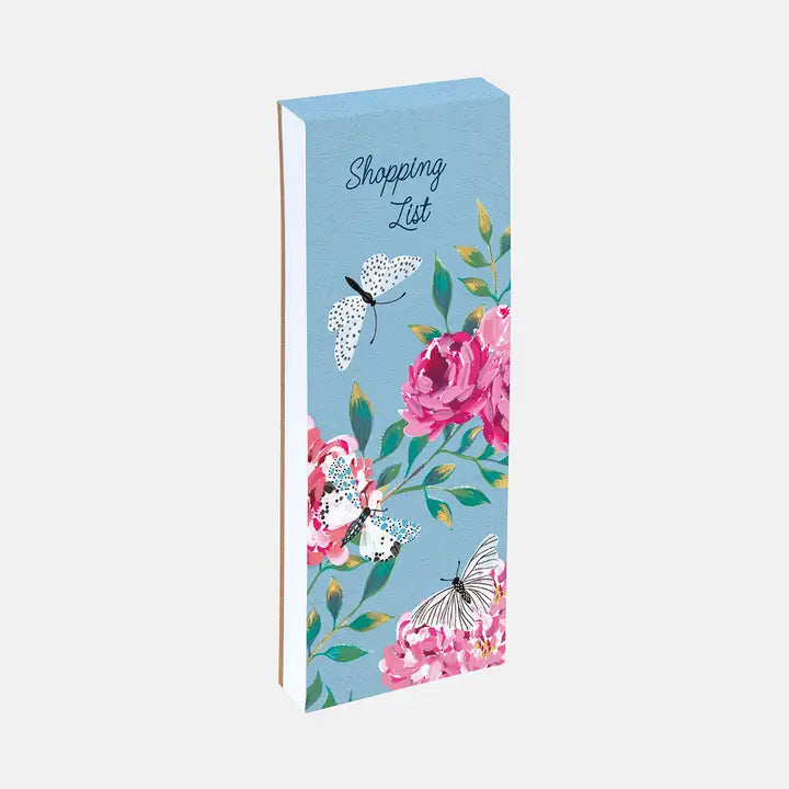 A tall, narrow shopping list notepad with a light blue cover decorated with pink flowers, green leaves, and white butterflies. The words 'Shopping List' are printed at the top.