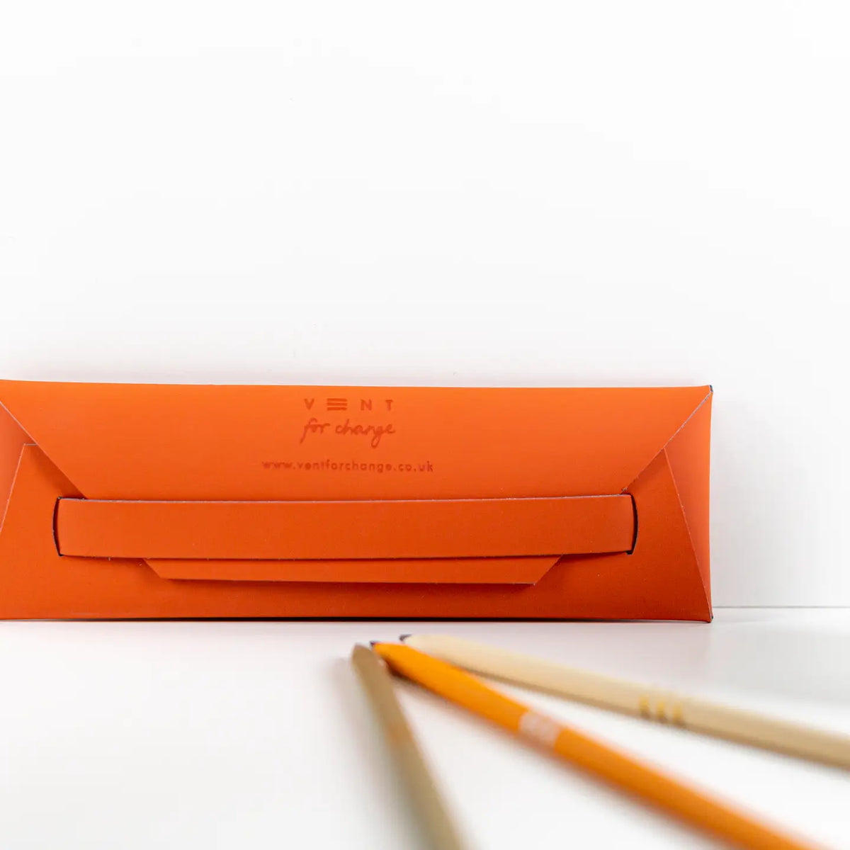 Back of an orange recycled leather pencil case showing its tuck-in closure, displayed beside pencils.